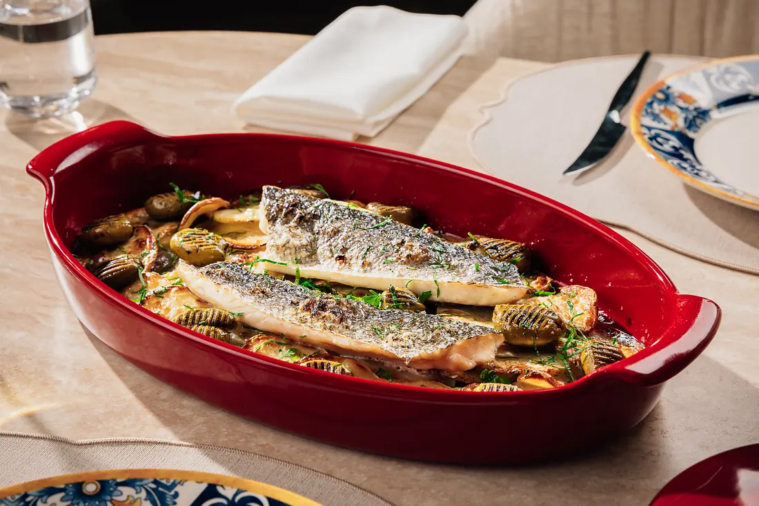 Two sea bass fillets baked with green olives, potatoes and herbs in a ceramic dish