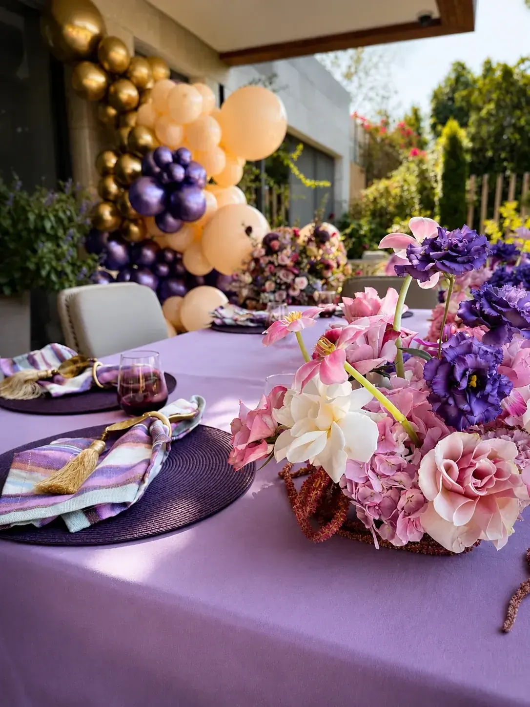 A celebration table in lilac linen with pink and purple florals and a balloon arch behind