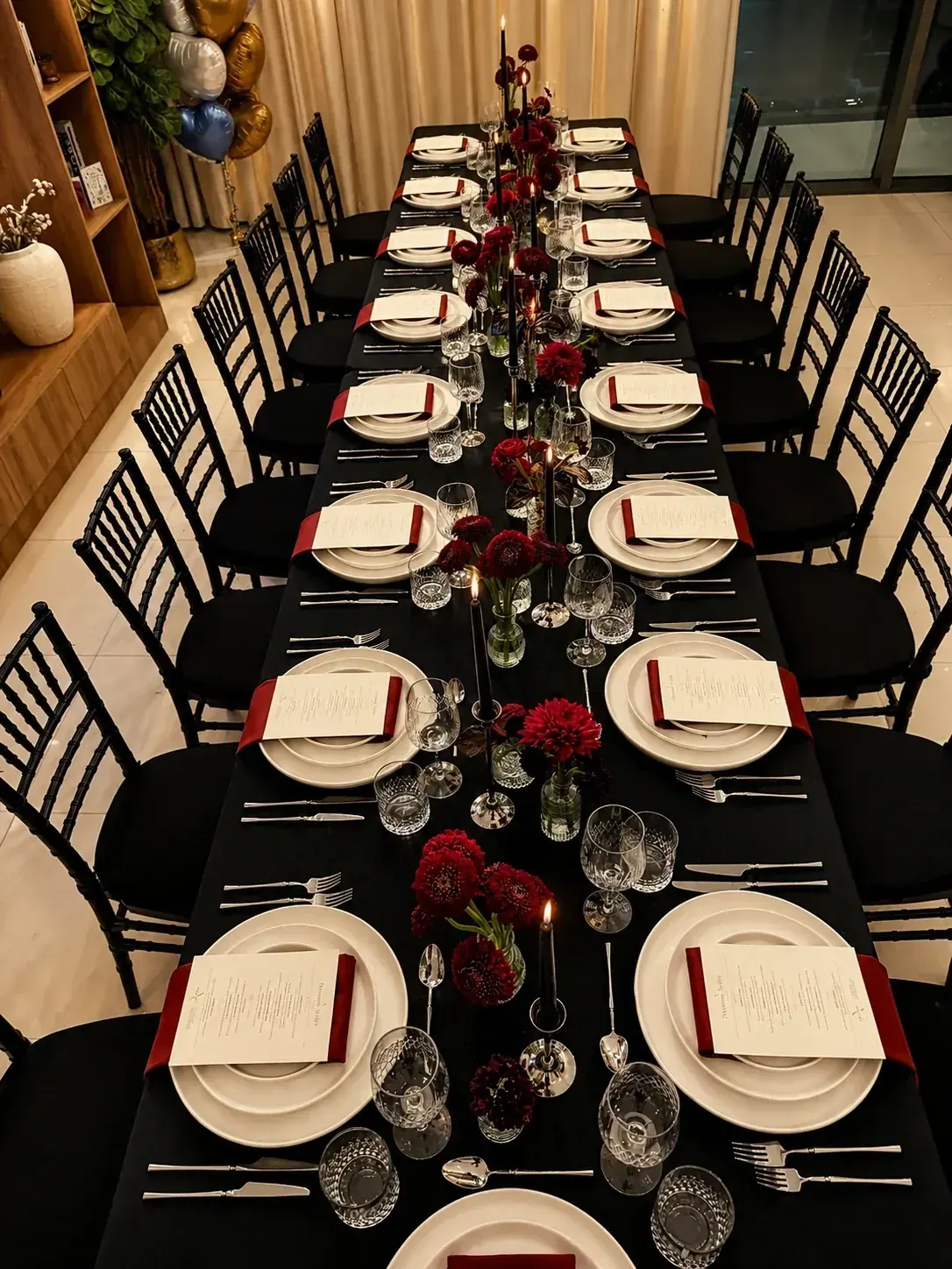 A long event table in black linen set for sixteen with red napkins, printed menus, tapered candles and red dahlias