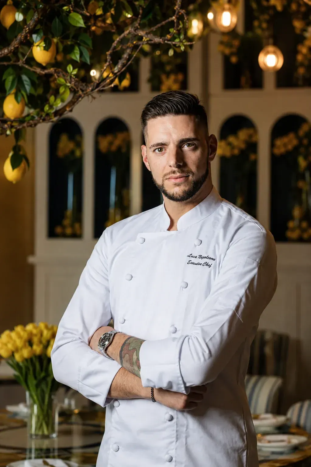 Chef Luca Napoleone in chef whites, arms folded, in a dining room dressed with lemon branches