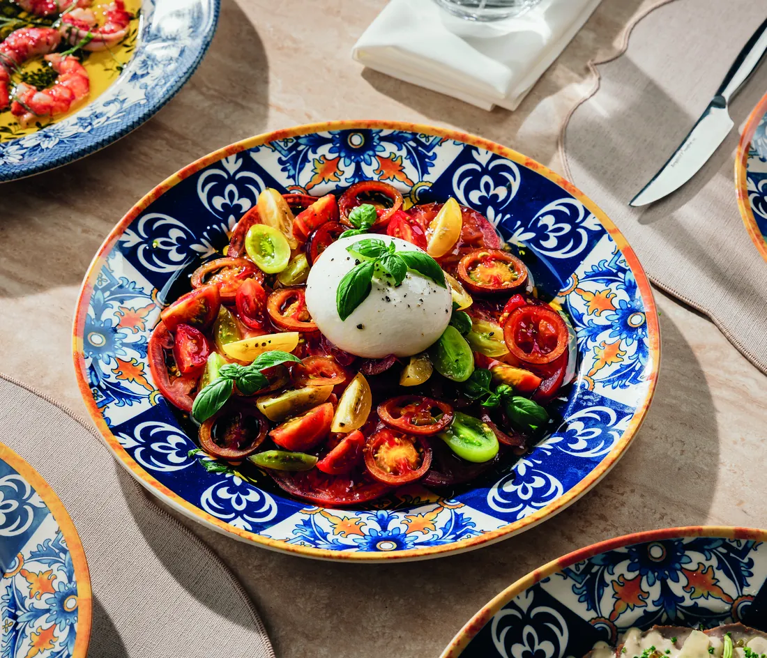 بوراتا مع طماطم من أصناف تراثية وريحان، في طبق مايوليكا مرسوم يدويًا، على مائدة مُعدّة