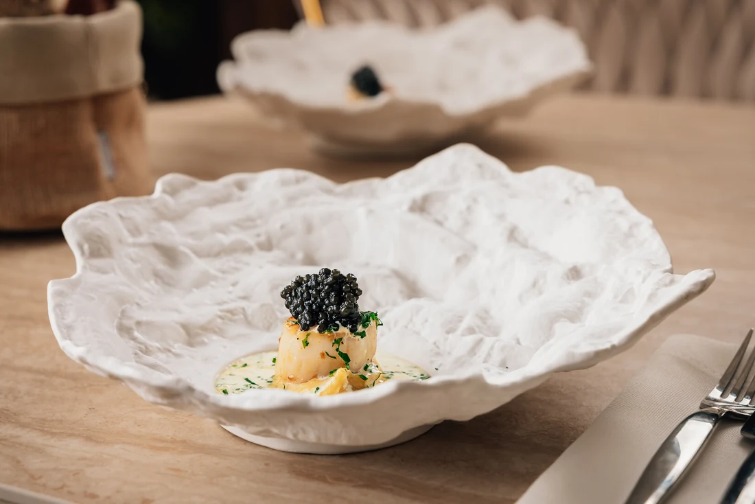 A seared scallop topped with caviar in a chive butter sauce, on a hand-sculpted white plate