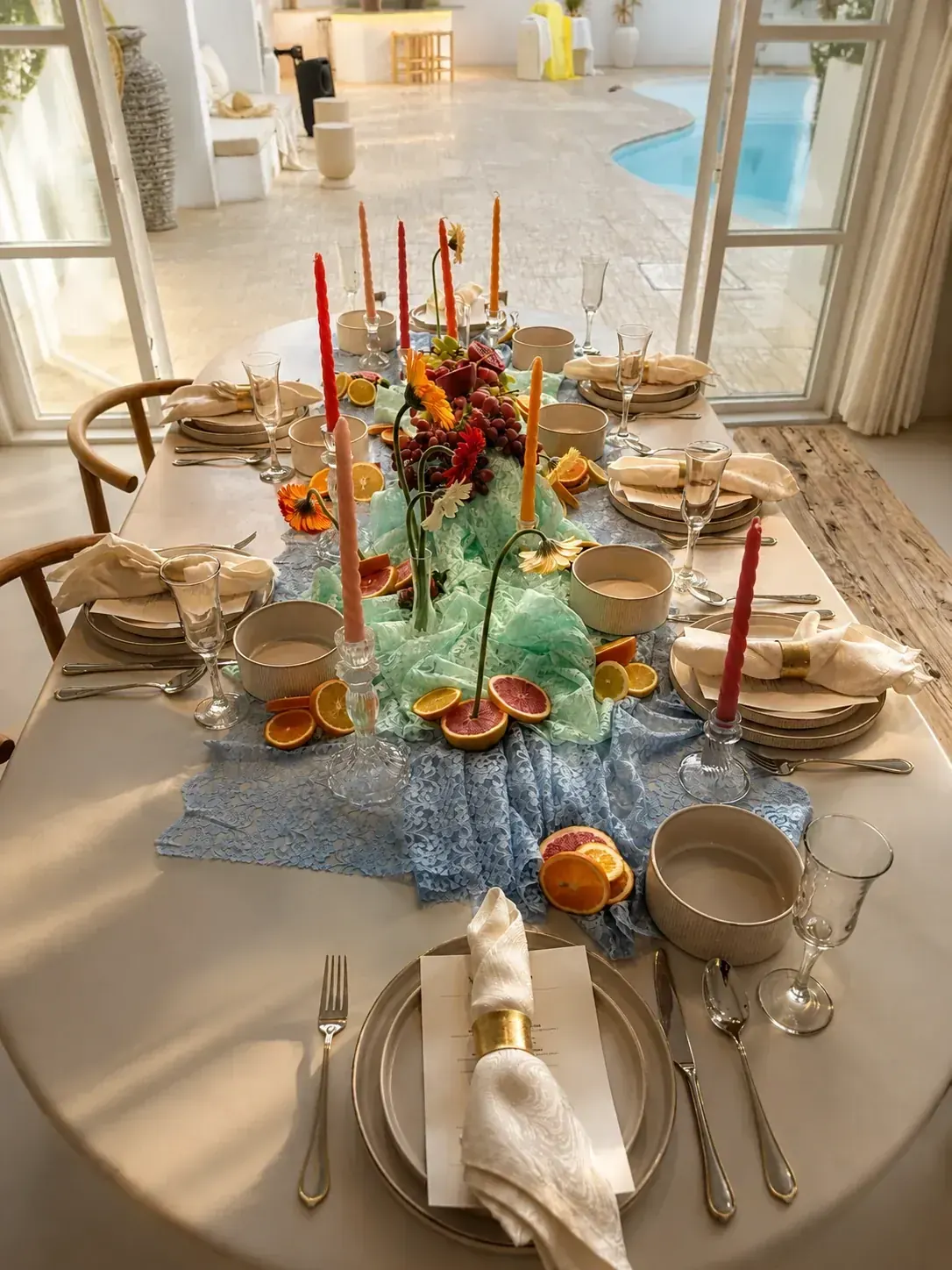 A villa dining table dressed in daylight with citrus, tapered candles and a lace runner, the pool visible beyond